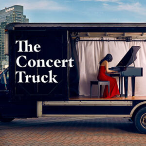 A black truck parked near a city with a woman in a red dress playing the piano with the words The Concert Truck