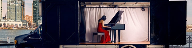 A black truck parked near a city with a woman in a red dress playing the piano with the words The Concert Truck